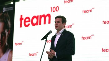 Minister Witold Bańka przemawia do mikrofonu, na tle logo Team100